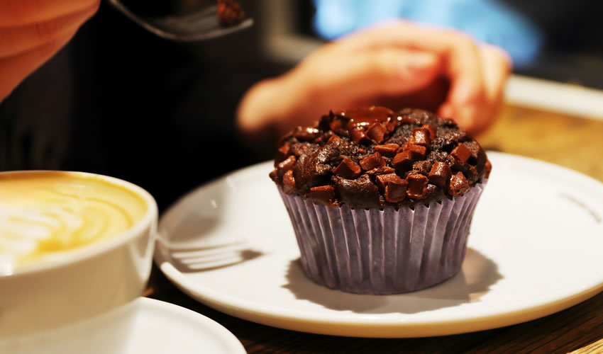 Chocolate muffin with chocolate chunks on a plate beside a latte on the table. Young womans hand holding a fork ready to taste delicious dessert.
