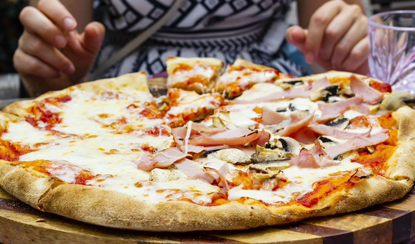 Large Italian pizza with mushrooms, ham, cheese on a wooden plate in the foreground with hands and body of a women in the background.